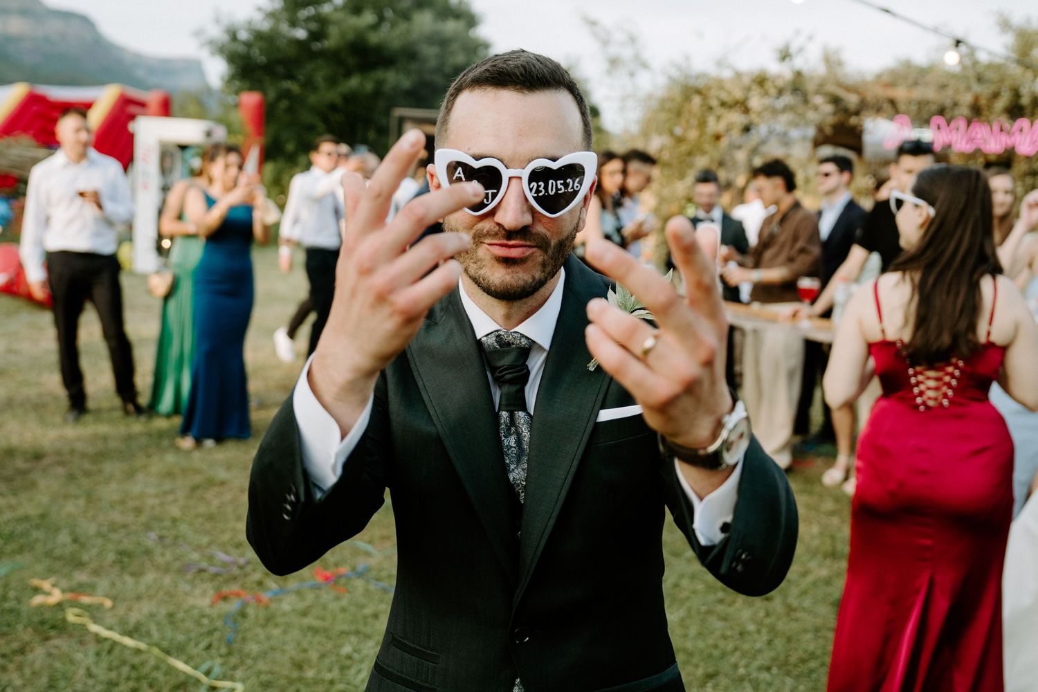 Groom in black suit wearing heart-shaped sunglasses at outdoor wedding reception, guests in formal attire in background.