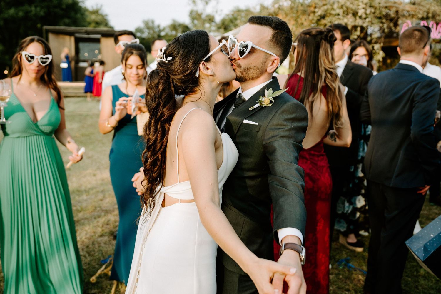 Bride and groom sharing a kiss at outdoor wedding reception with guests in elegant attire in background.