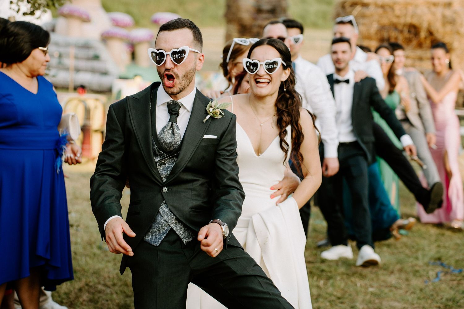 Bride and groom dancing joyfully in heart-shaped sunglasses at outdoor wedding celebration with guests.