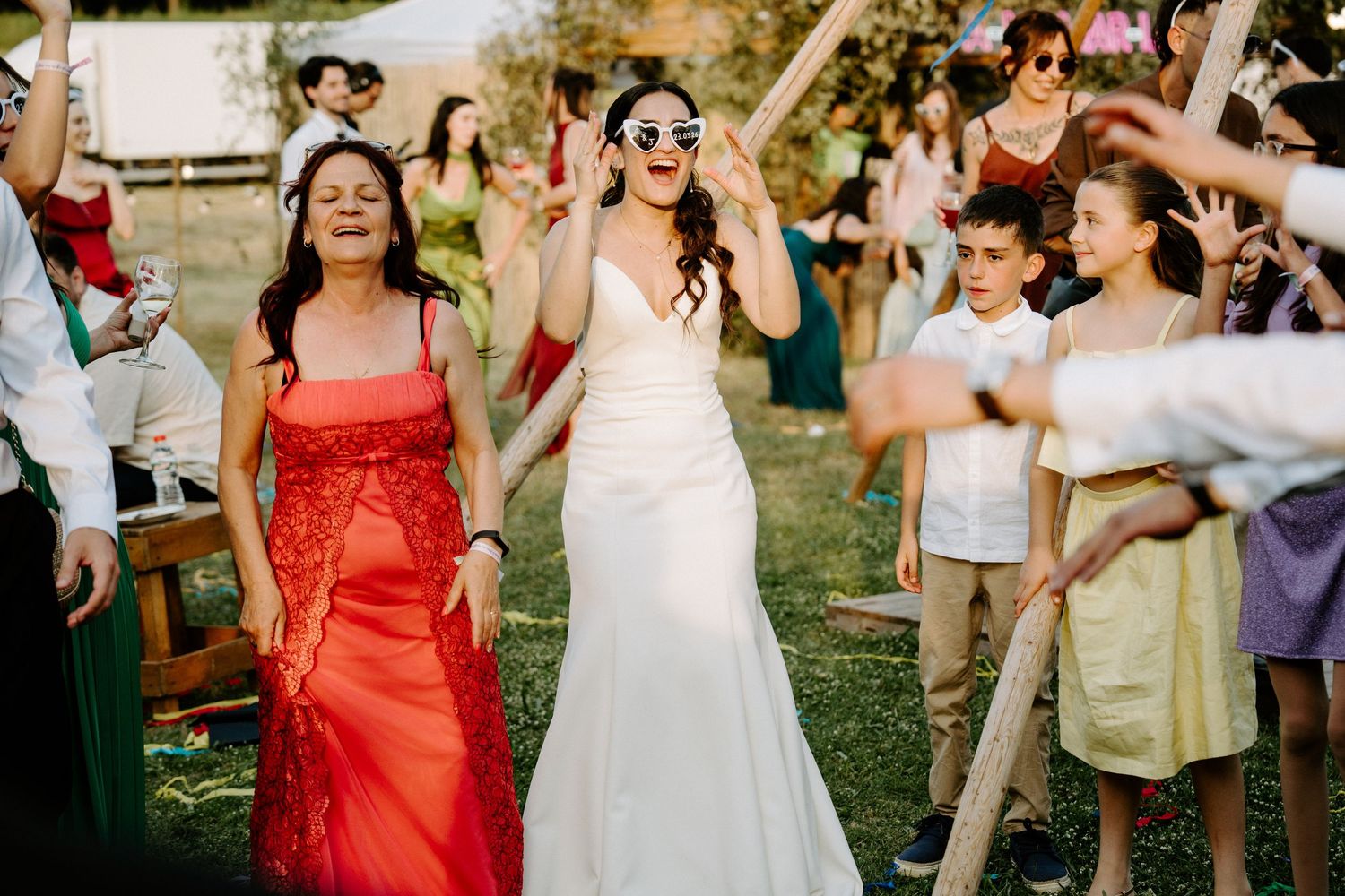 Bride in white gown walks with guests at outdoor wedding celebration, wearing sunglasses while crowd cheers.