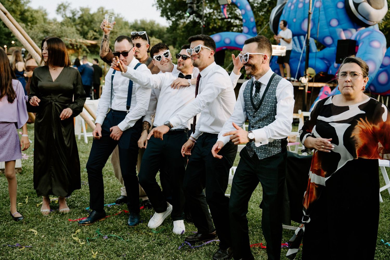Groomsmen in suspenders and sunglasses dancing outdoors at a wedding reception with colorful decorations.
