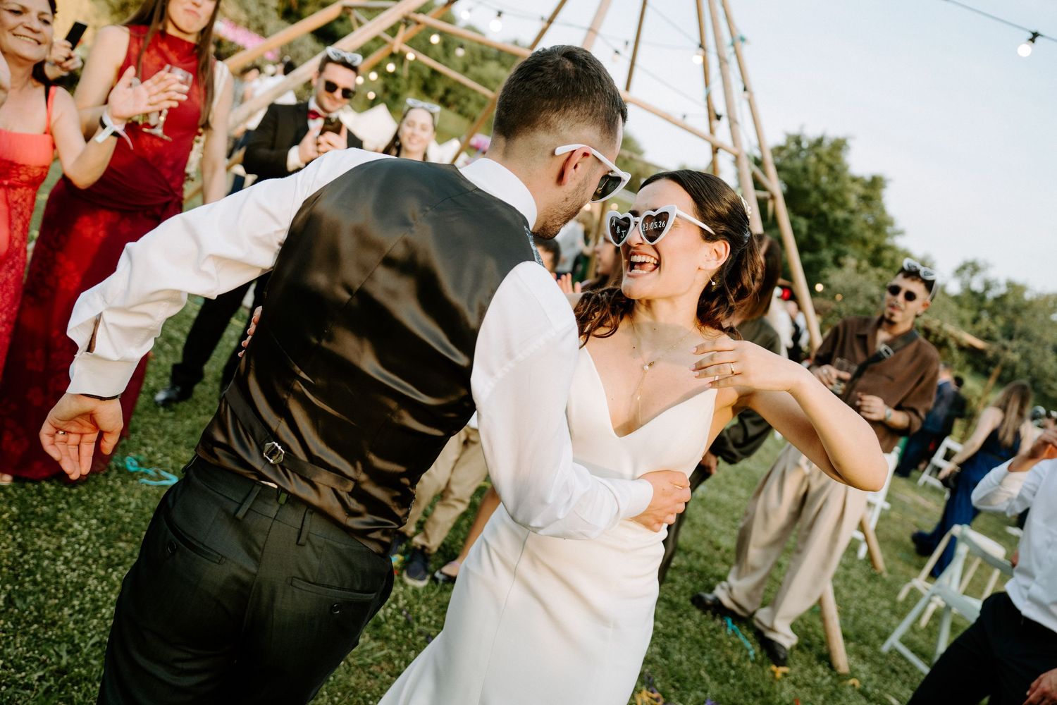 Bride and groom dancing joyfully outdoors at wedding reception wearing sunglasses under string lights.
