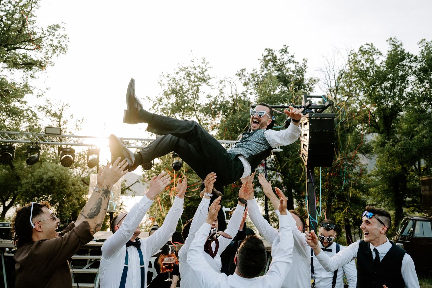 Groomsmen tossing groom into the air in celebration at outdoor wedding reception near stage.