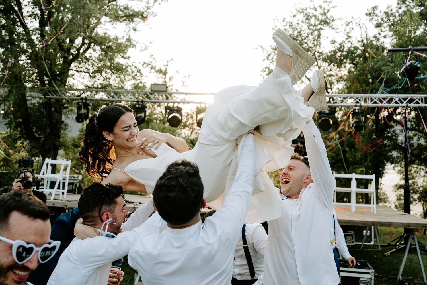 Joyful wedding guests lifting the smiling bride into the air during an outdoor wedding celebration.