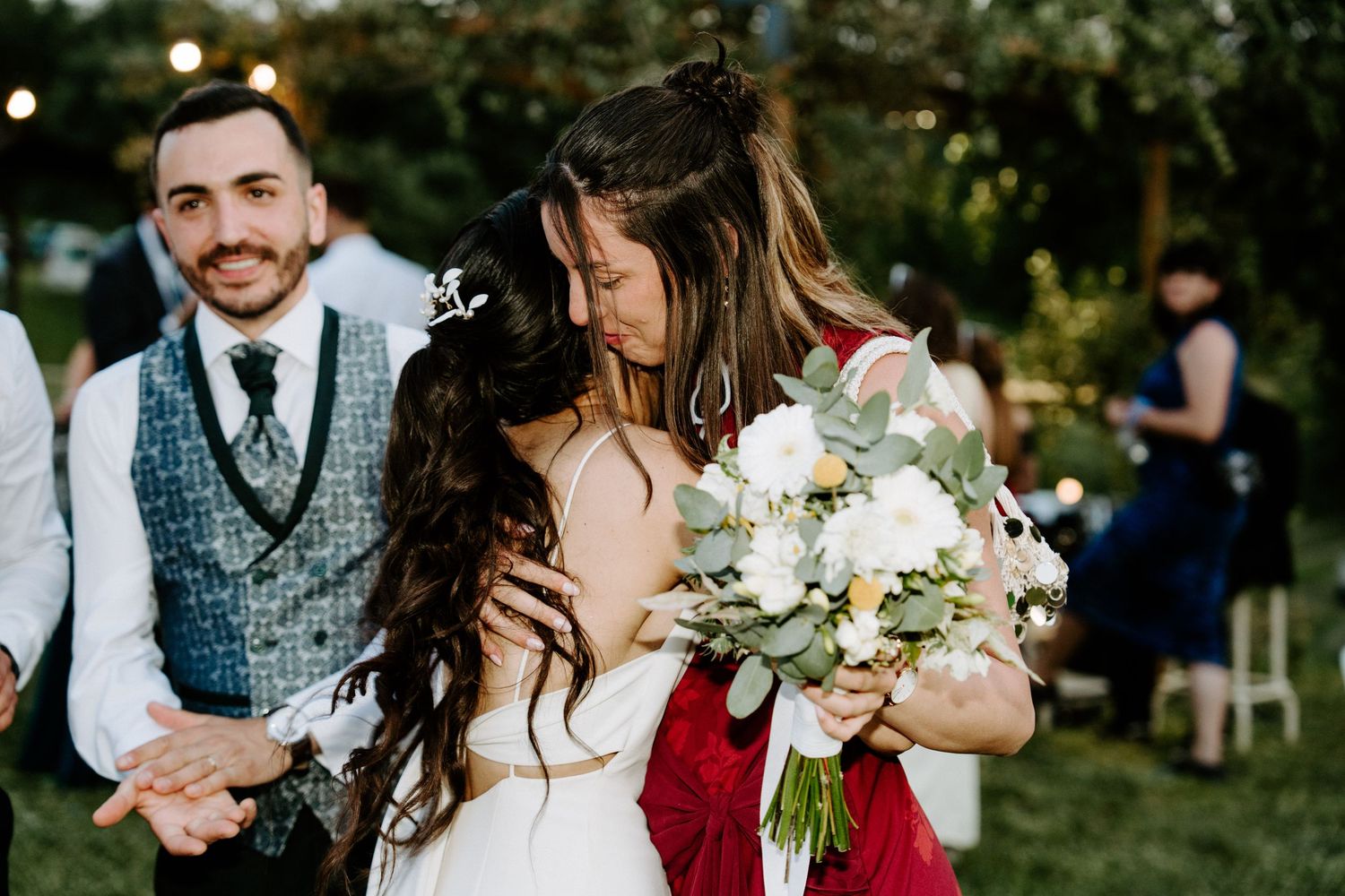 Bride hugging a guest in a red dress while holding a bouquet at an outdoor wedding ceremony.