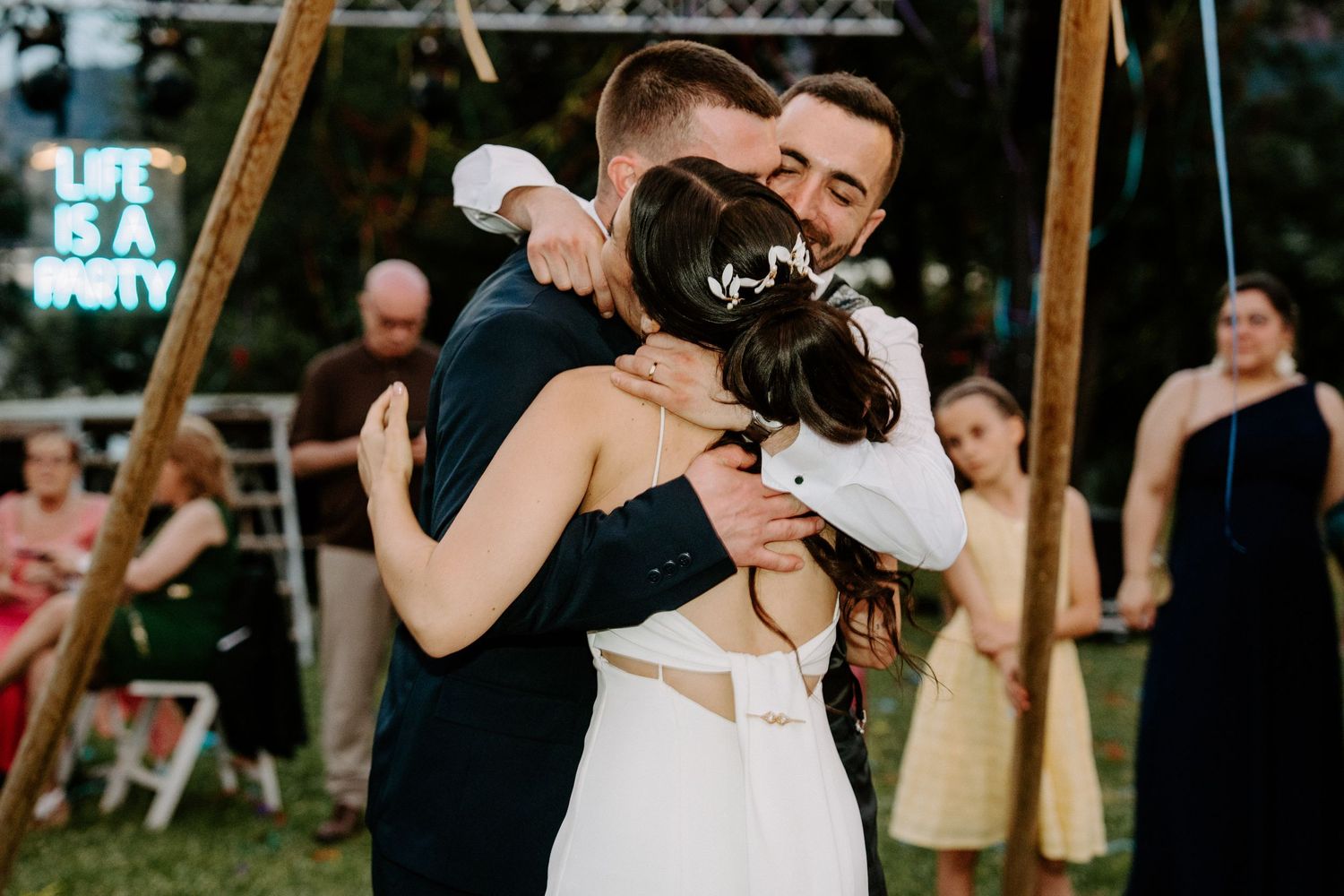 Bride and groom share a heartfelt group hug with a guest at an outdoor wedding reception.