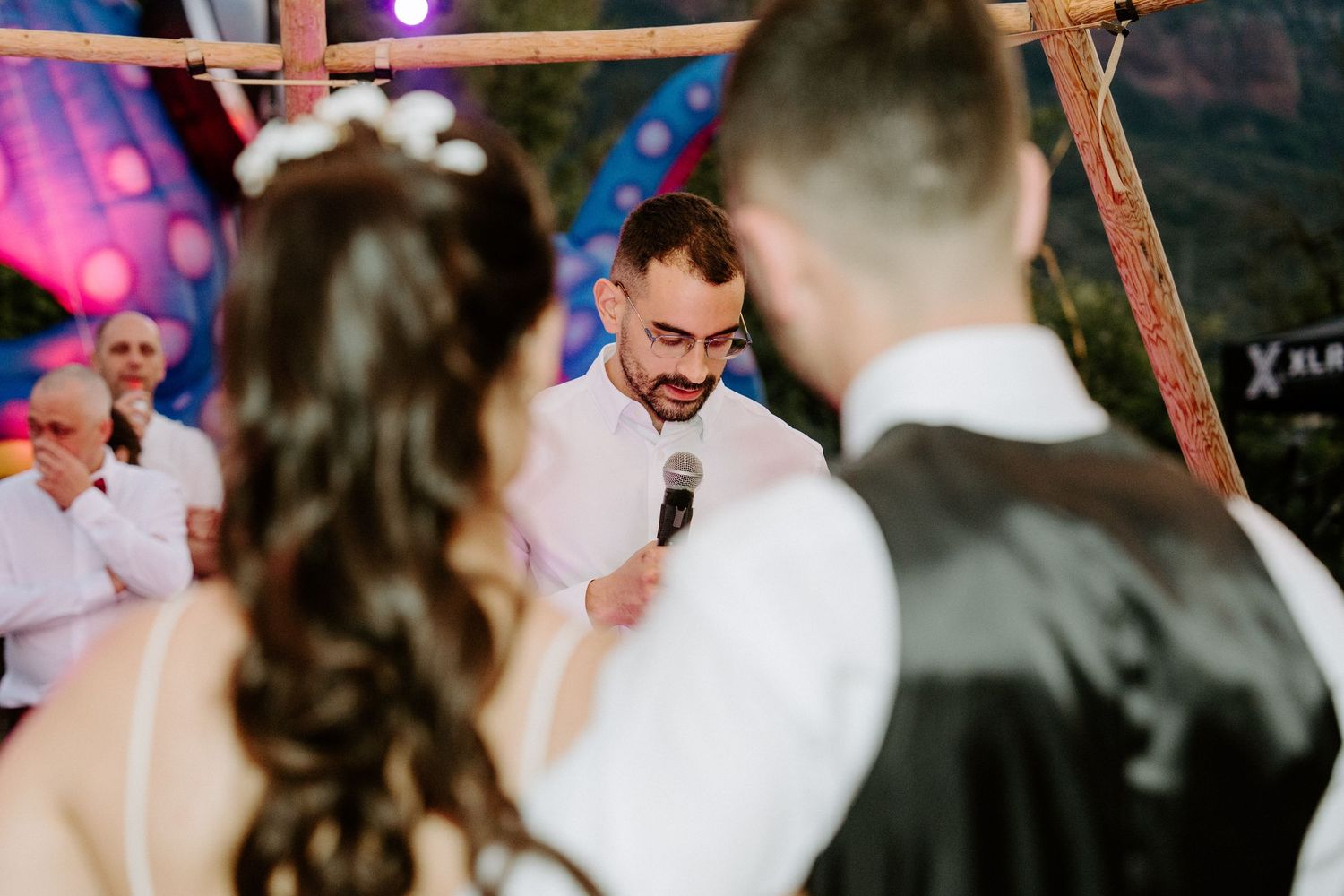 Groom speaks into microphone at outdoor wedding reception with colorful lights in background.