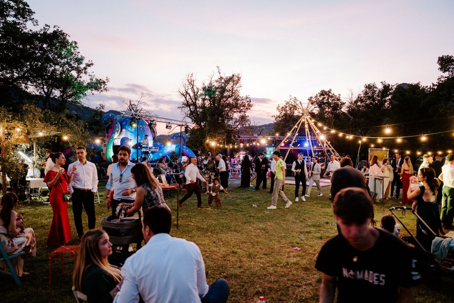 Outdoor evening wedding reception with string lights, tipi tent, and guests mingling on a lawn at dusk.