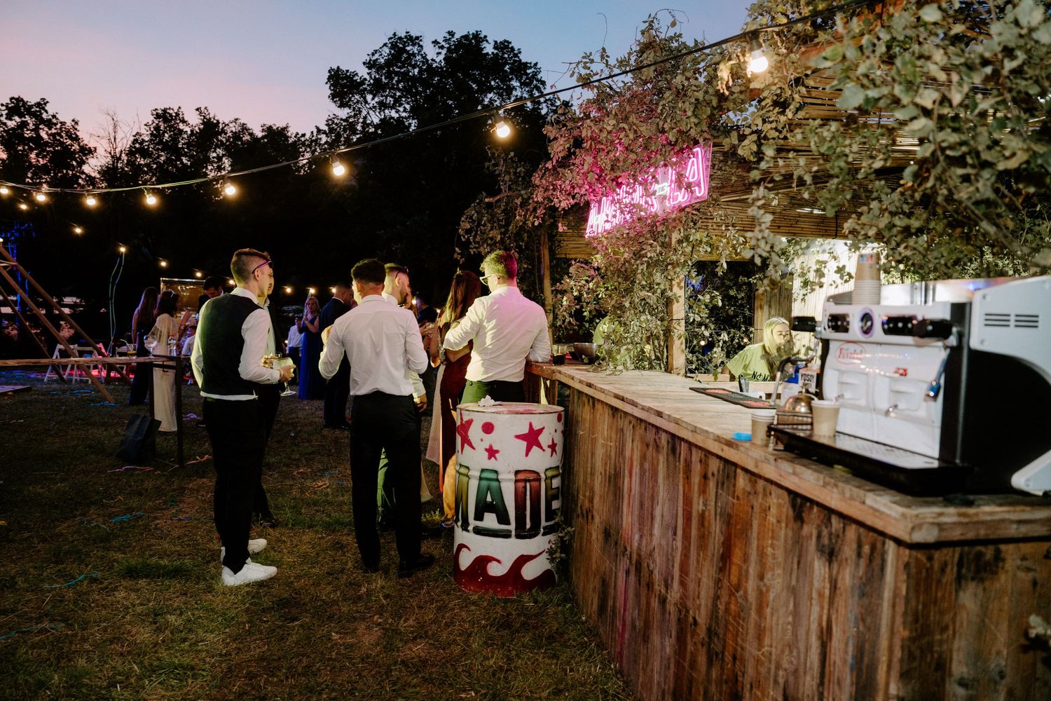Guests gather at a rustic outdoor bar with string lights at a twilight wedding reception.