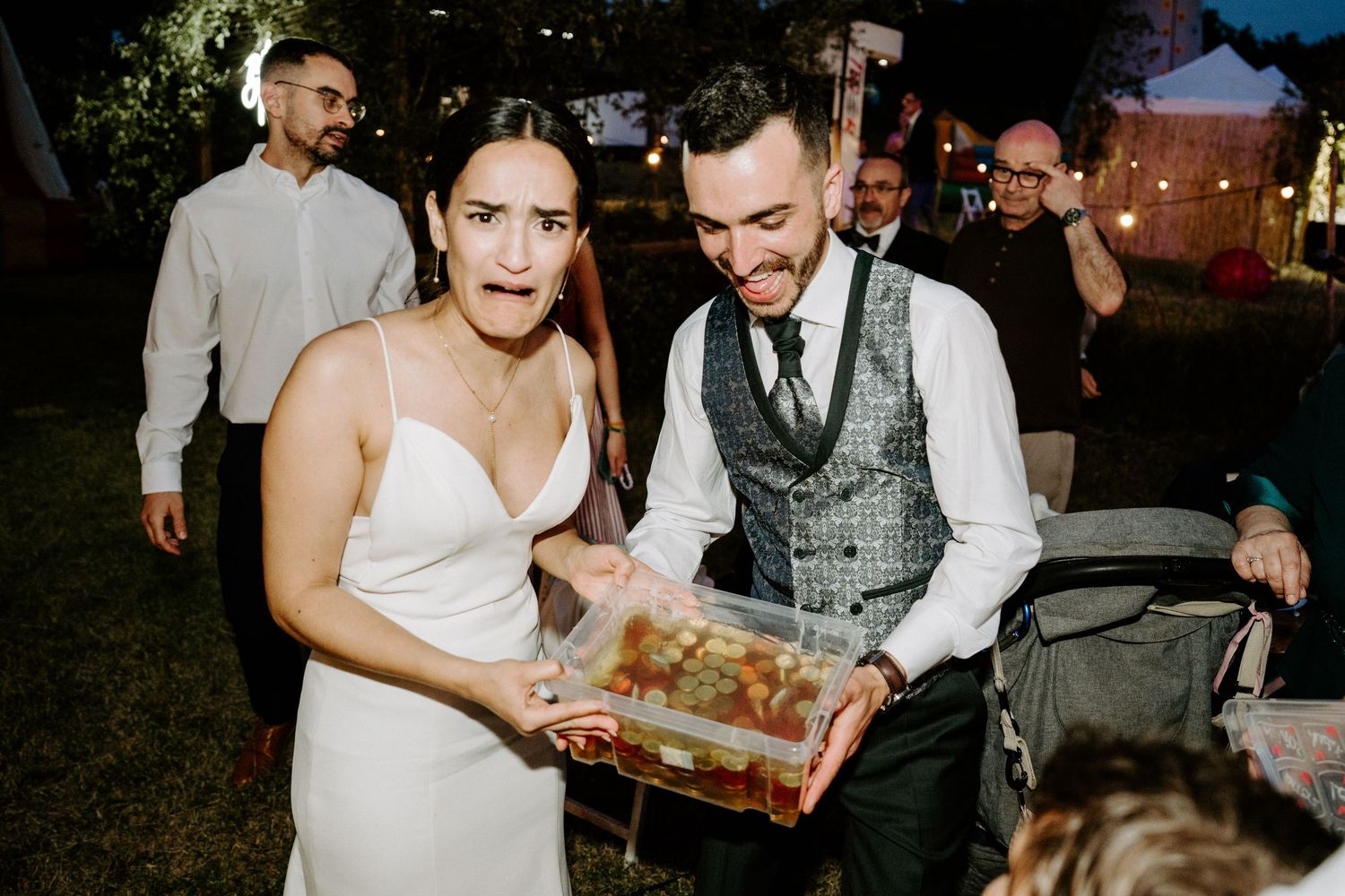 Bride and groom smiling while holding a clear box of treats at their outdoor evening wedding reception.