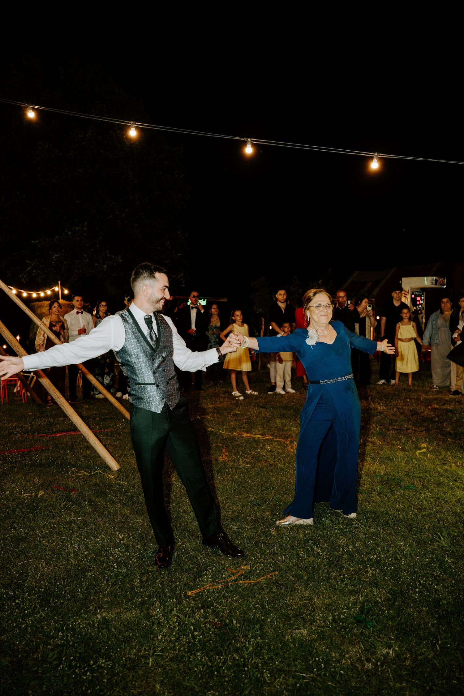 A groom and female guest dance joyfully outdoors at a night wedding reception with string lights.
