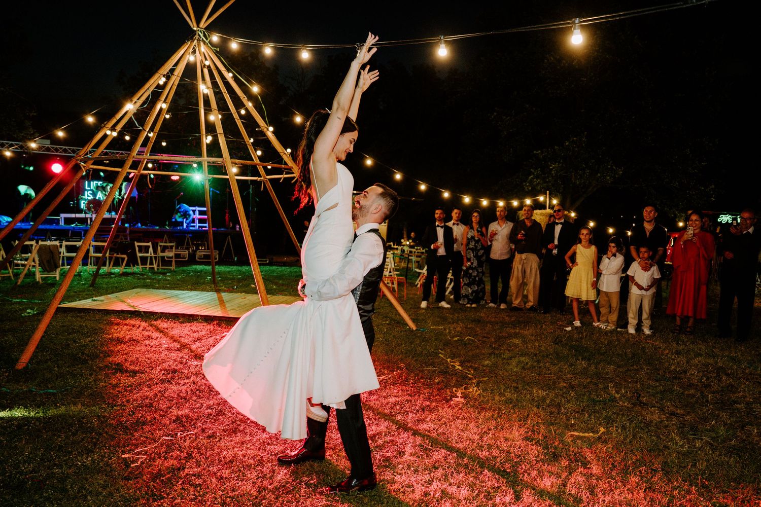 Bride and groom share a romantic first dance under string lights near a tipi at an outdoor wedding reception.