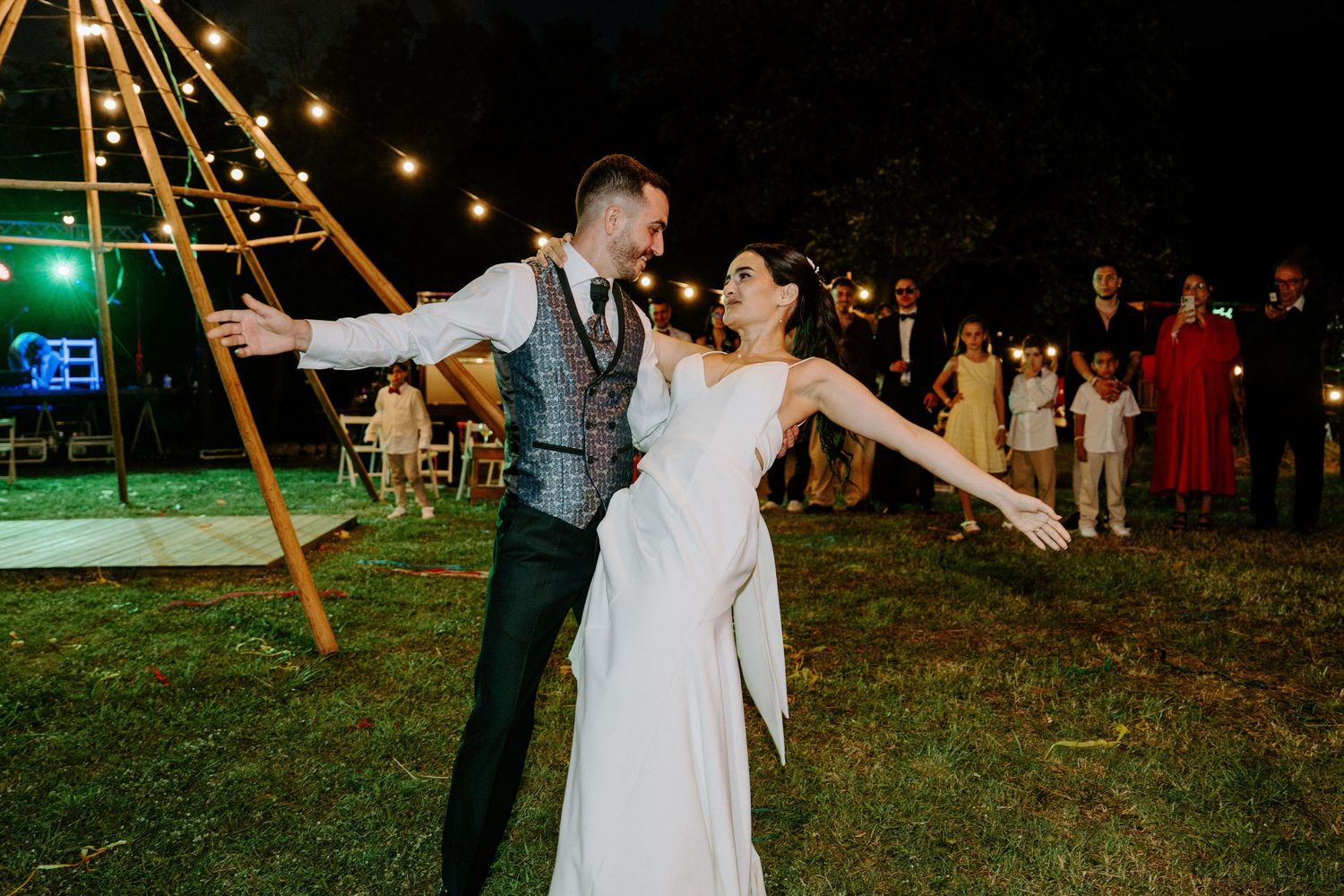 Bride and groom share a joyful first dance on the grass at an outdoor evening wedding.