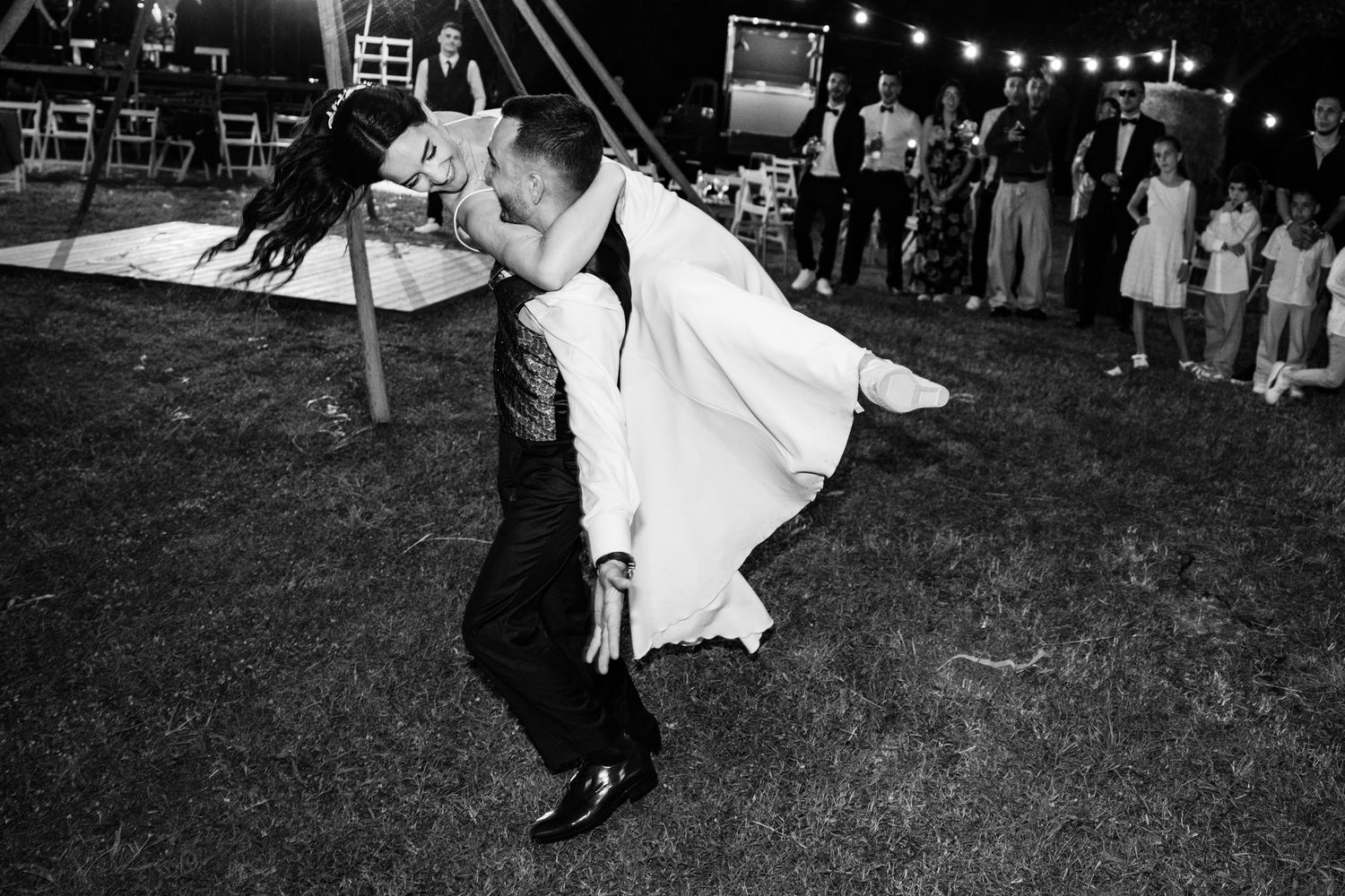 Black and white photo of groom dipping bride during wedding first dance outdoors under string lights.