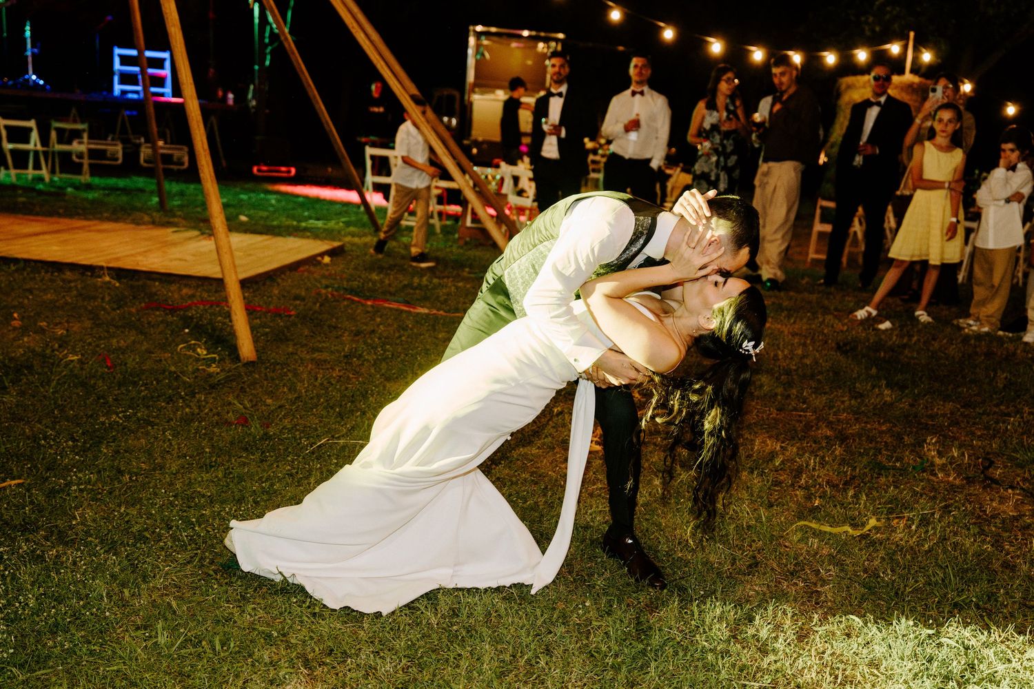 Groom dips bride for a romantic kiss on the grass at an outdoor wedding reception at night.