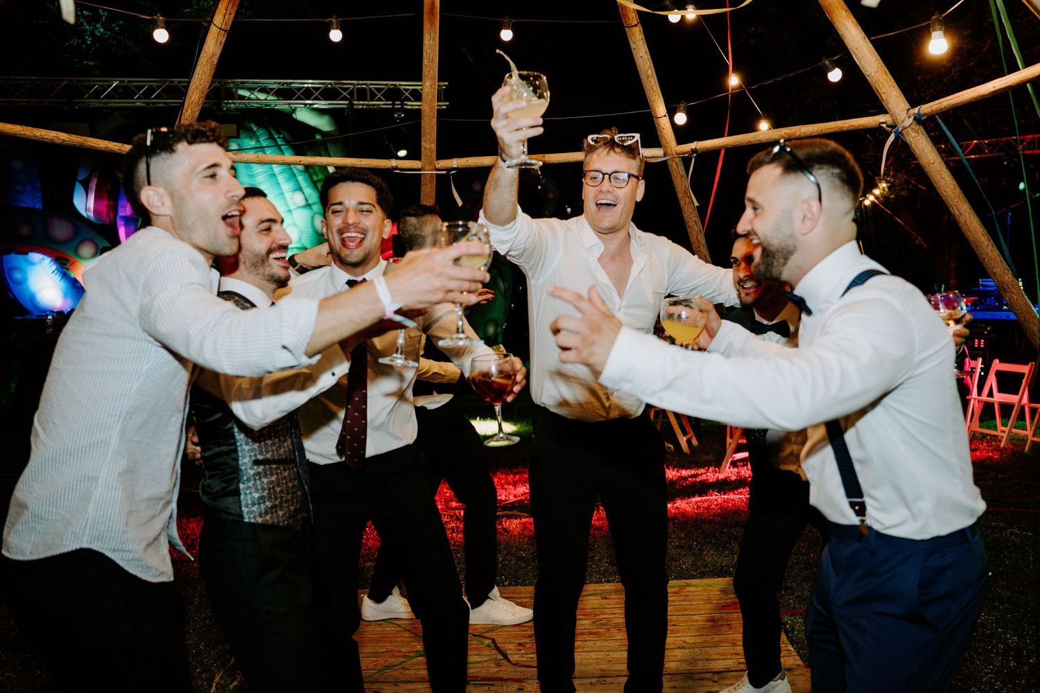 Groomsmen celebrating and toasting drinks together inside a tipi at a lively wedding reception party.