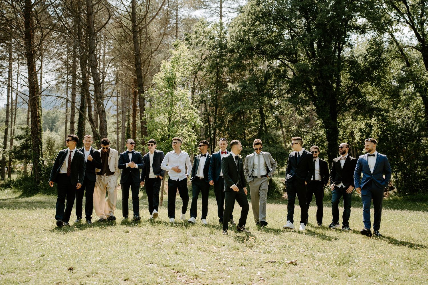 Groomsmen in suits laughing and posing together outdoors in a forest clearing on a wedding day.