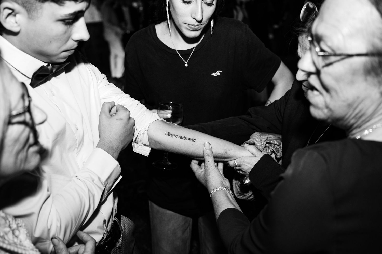 Black and white photo of wedding guests admiring a tattoo on a young mans arm at reception.