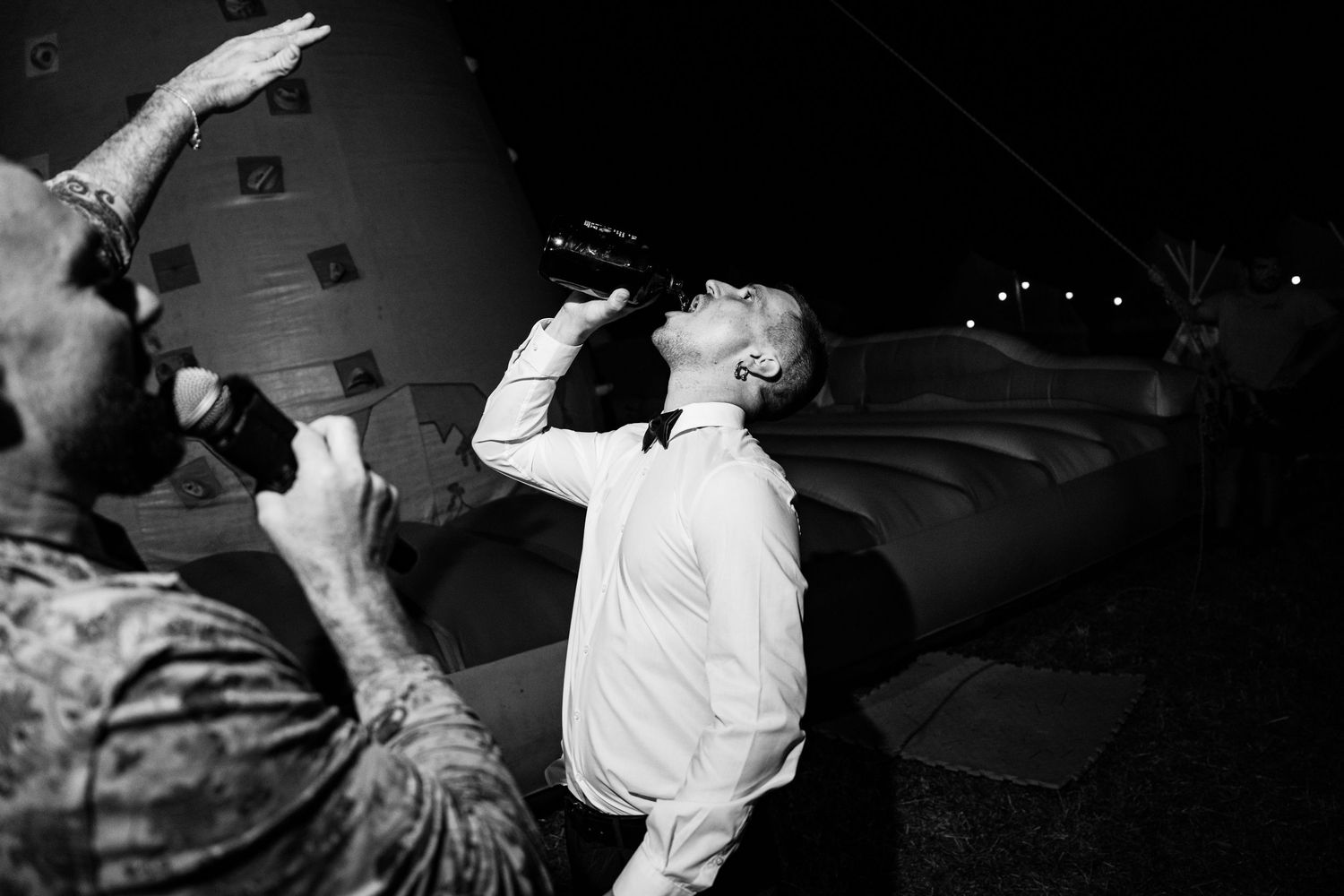 Black and white photo of man drinking from bottle at wedding reception.