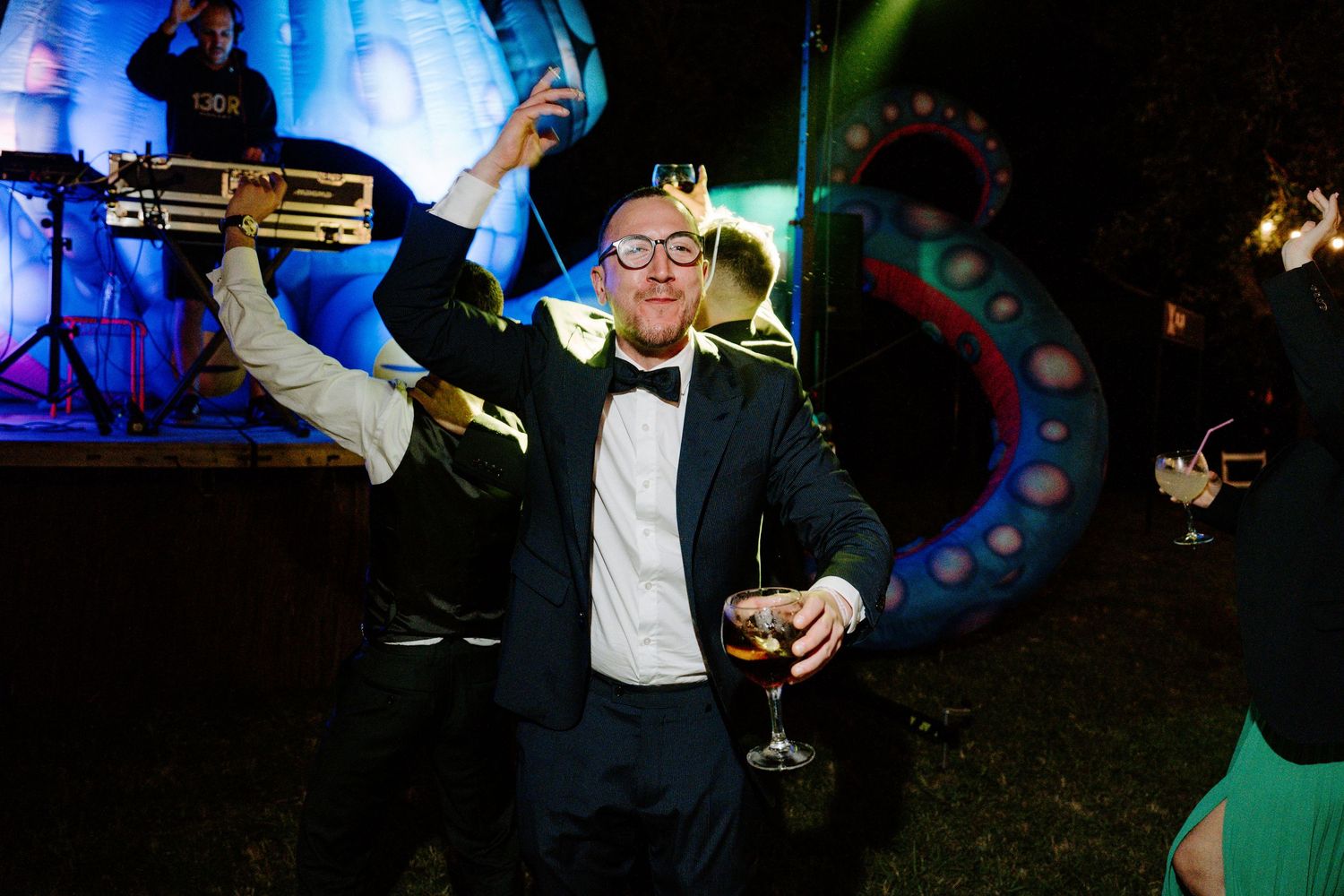 A man in a tuxedo celebrates at a wedding party, holding a champagne glass with a live band behind him.