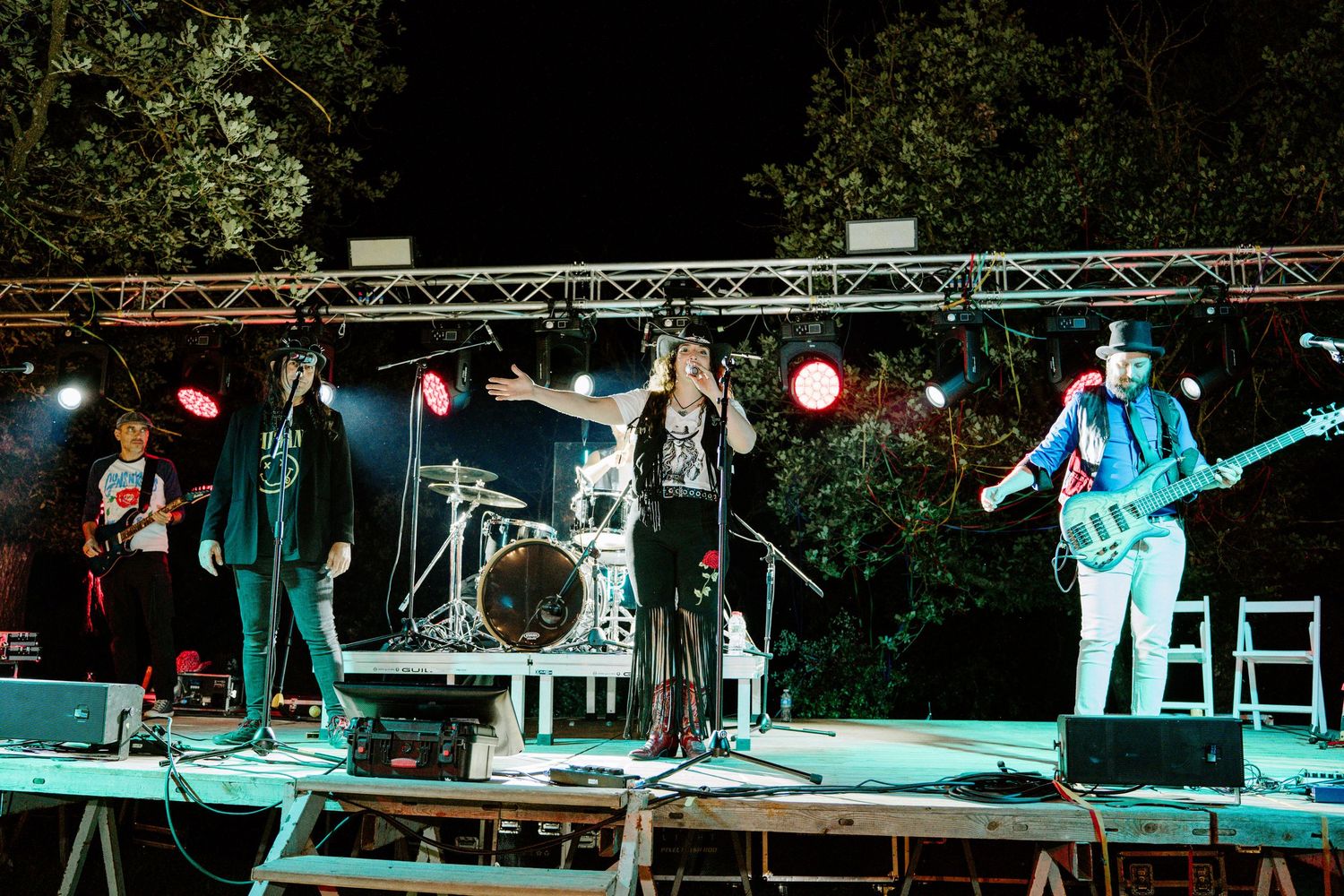 Rock band performing on an outdoor stage at night with colorful lights and trees in the background.