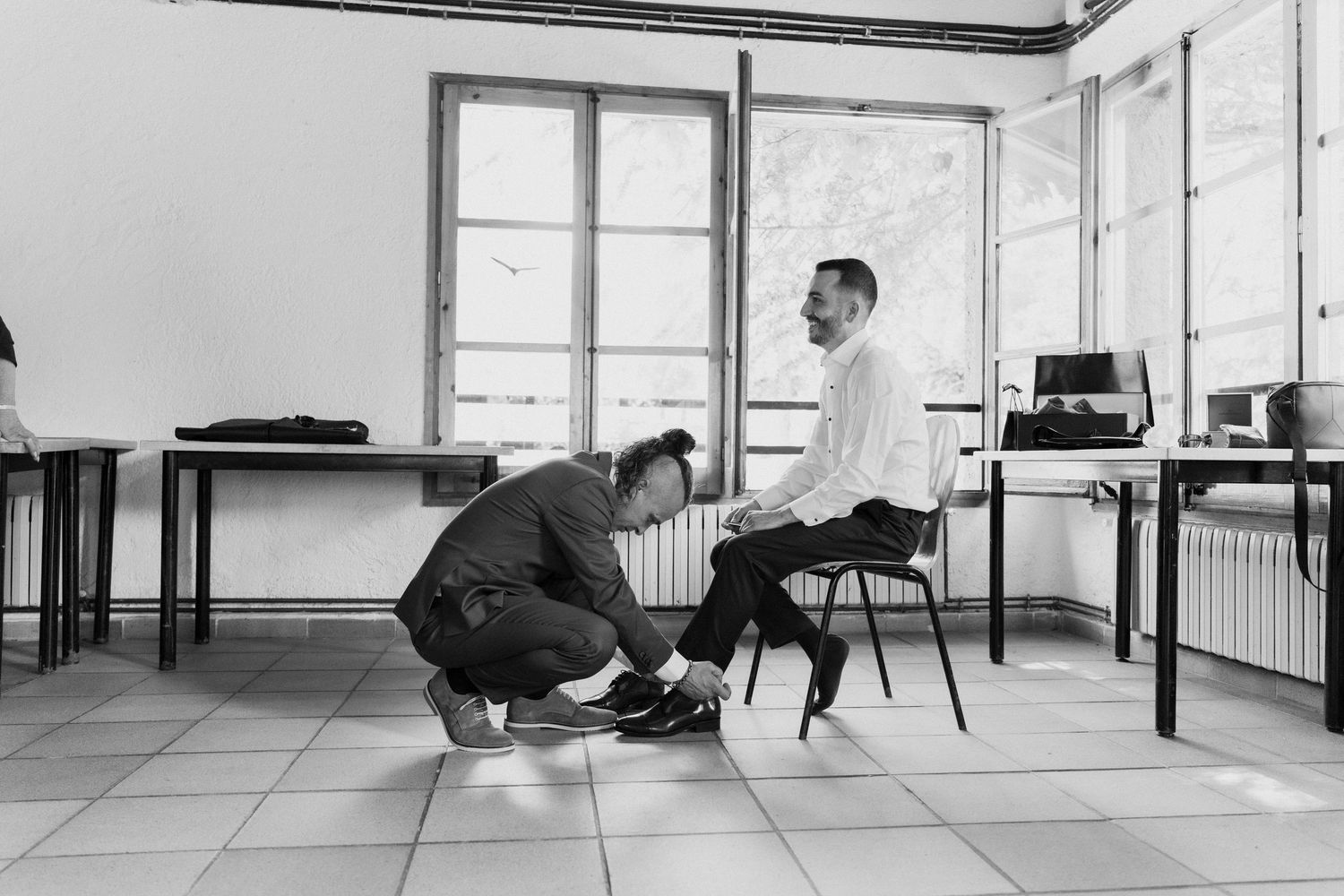 Black and white photo of groomsman helping groom with shoes in preparation room before wedding.