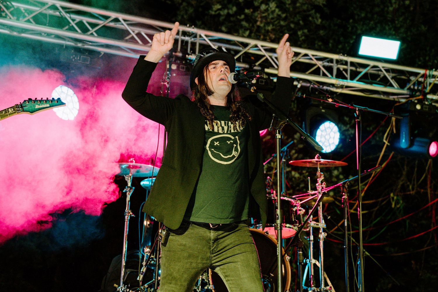Energetic performer raises hands on stage with pink smoke and lights at an outdoor concert.
