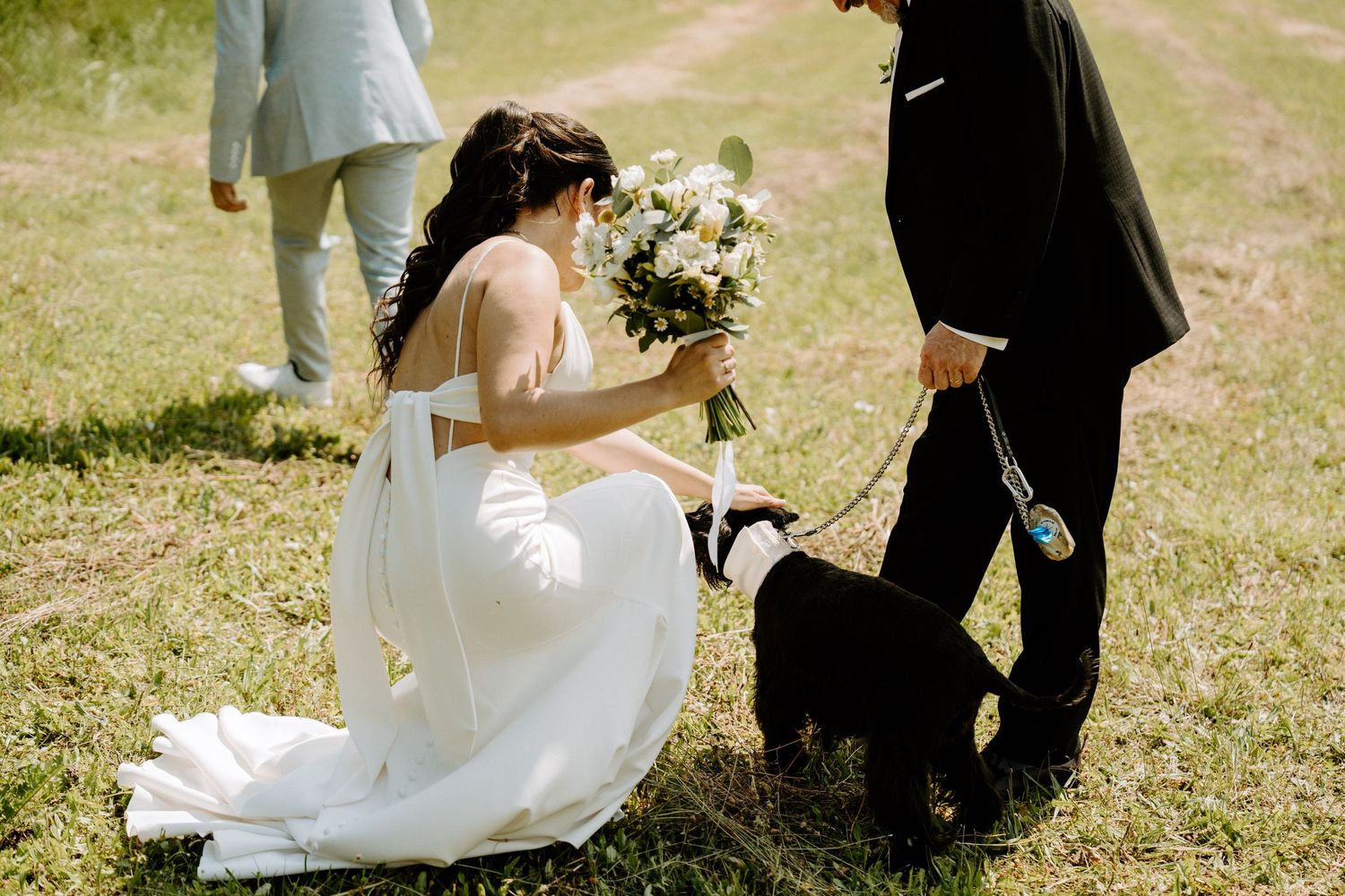 A bride in a white gown kneels to greet a black dog while holding a bouquet at an outdoor wedding.