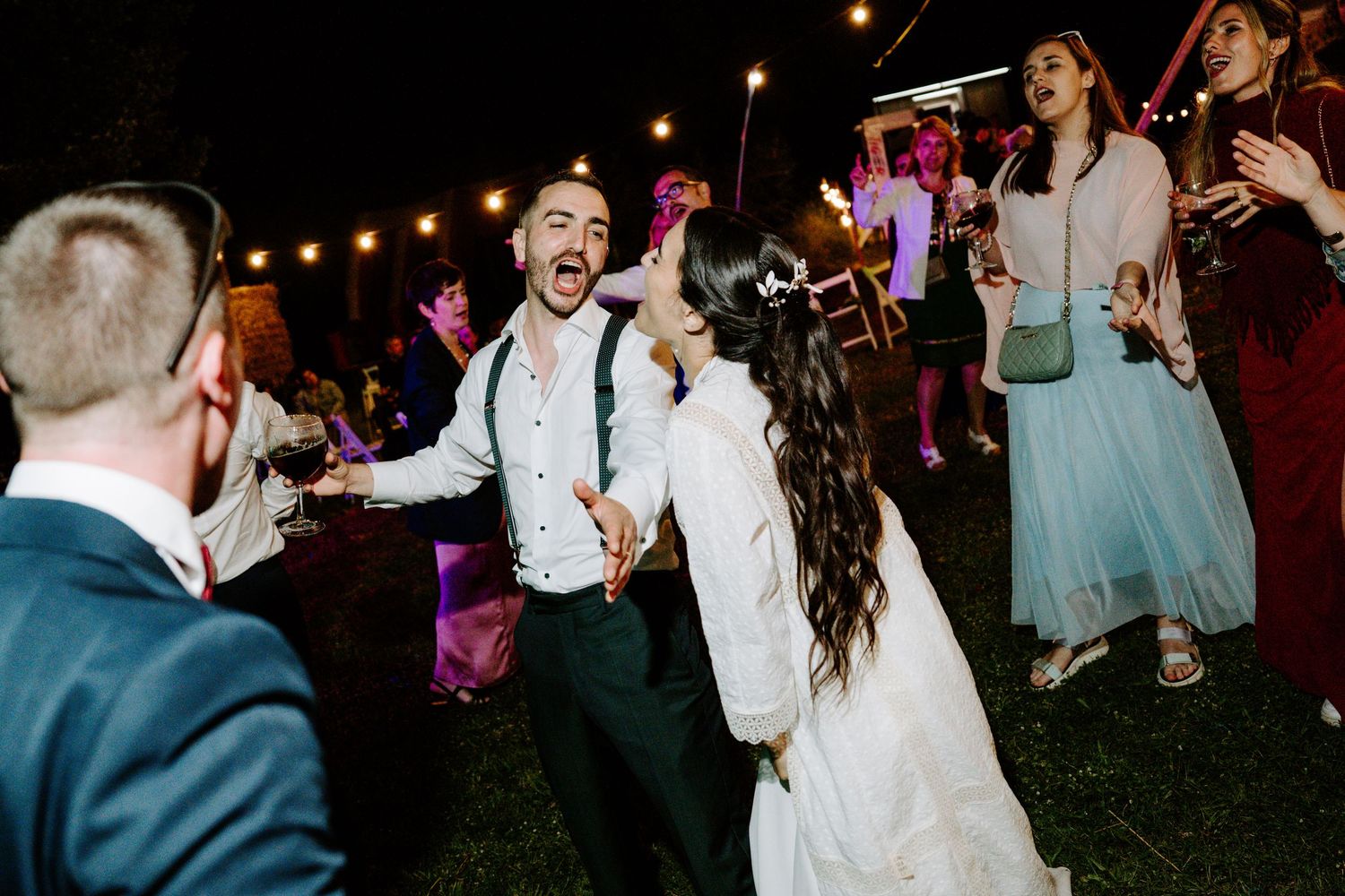 Bride and groom dancing and laughing together with guests at a lively outdoor evening wedding reception.