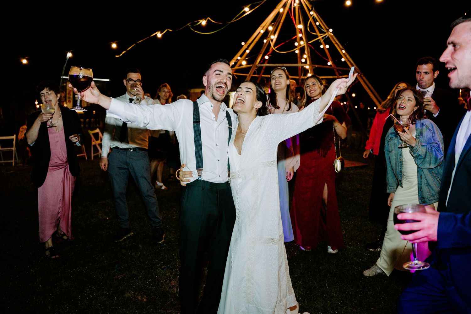 Joyful bride and groom celebrate with arms raised at festive outdoor wedding reception at night.