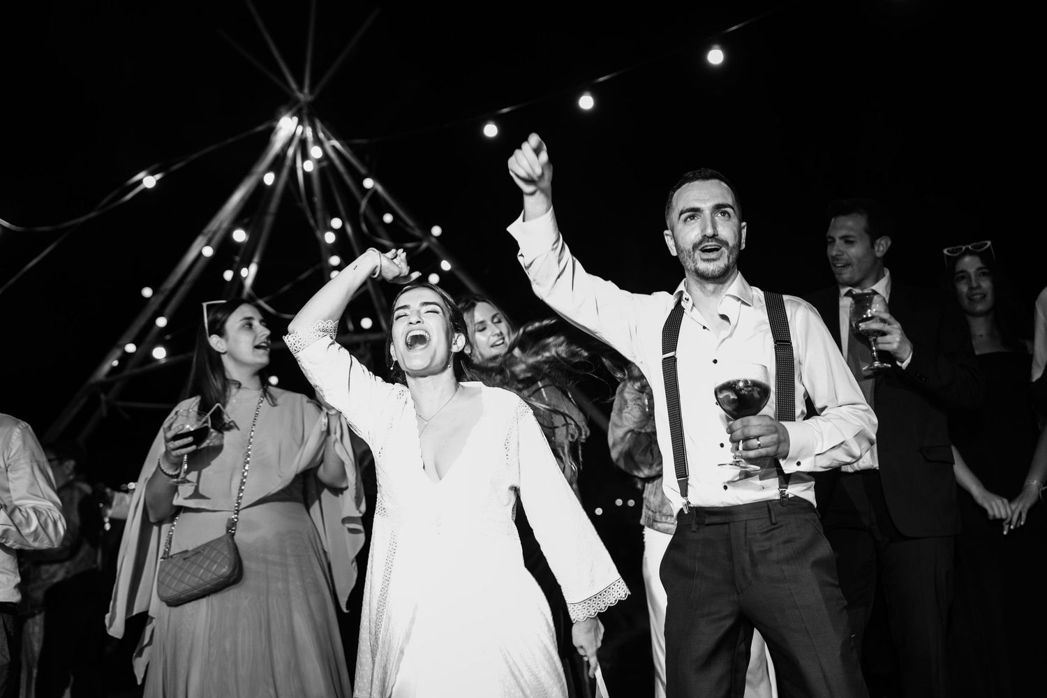 Black and white photo of bride and groom celebrating joyfully under string lights at wedding reception.