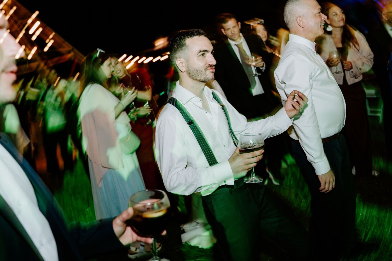 Guests dancing and socializing at an outdoor evening wedding reception, men wearing white shirts with green suspenders.