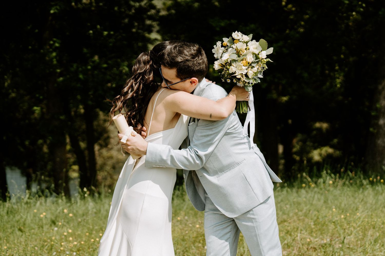 Bride and groom embracing in green meadow, bride holding white floral bouquet.