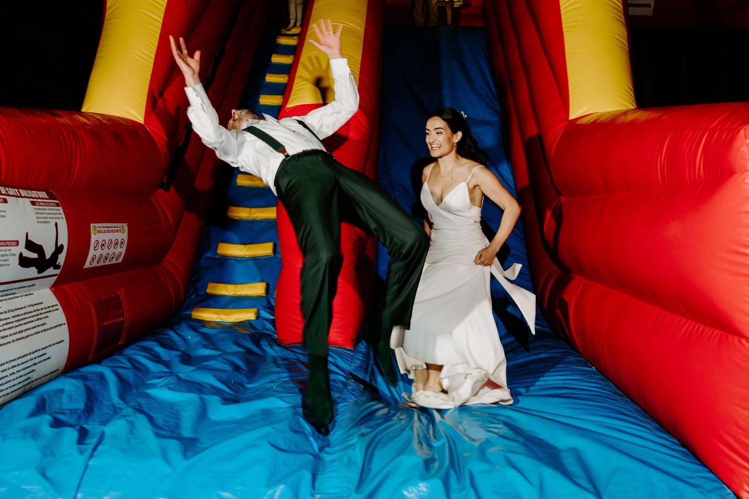 Bride and groom joyfully jumping on a colorful inflatable bounce house in wedding attire.