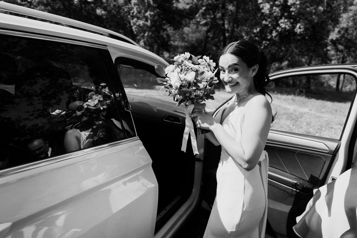 Smiling bride in white gown holding bouquet stepping out of a vintage car in black and white photo.