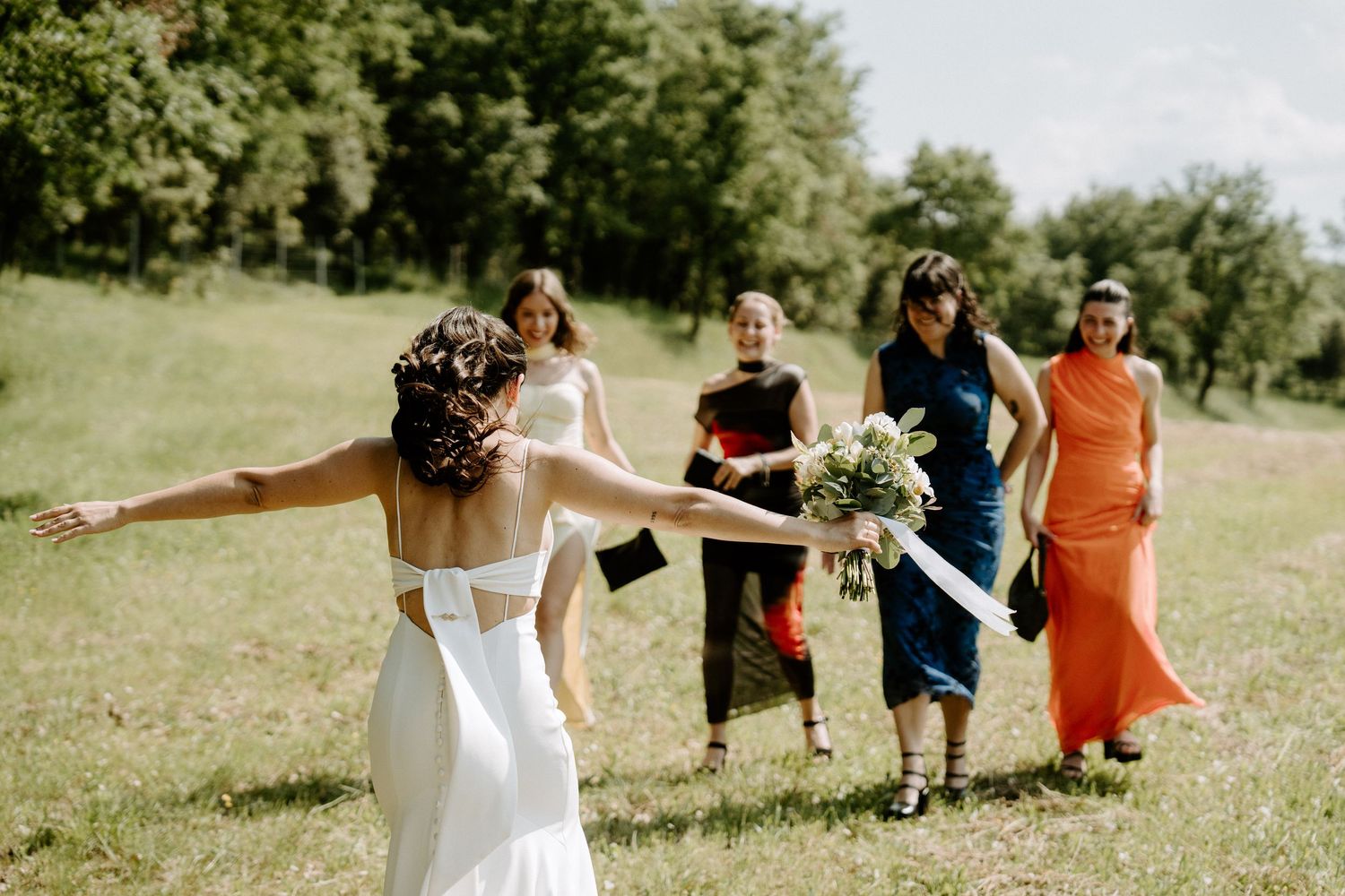 Bride in backless white gown with arms outstretched greeting bridesmaids in colorful dresses outdoors.