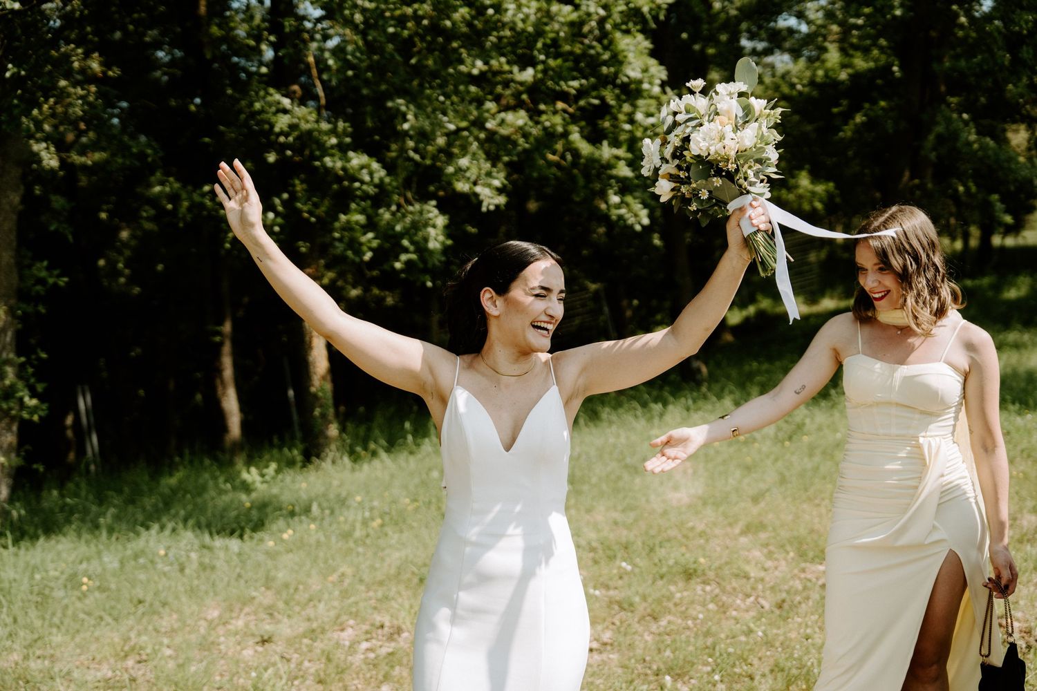 Joyful bride raises arms holding bouquet while walking with bridesmaid outdoors.