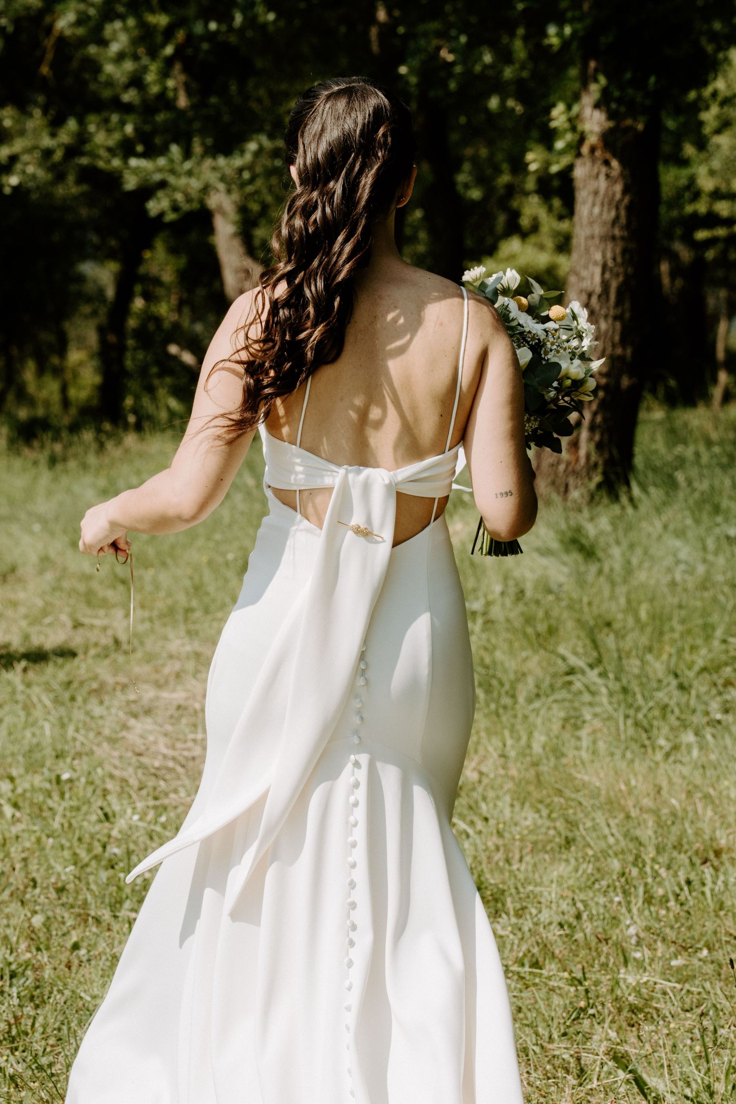 Bride in backless white satin gown with bow detail holding bouquet walking through green forest.