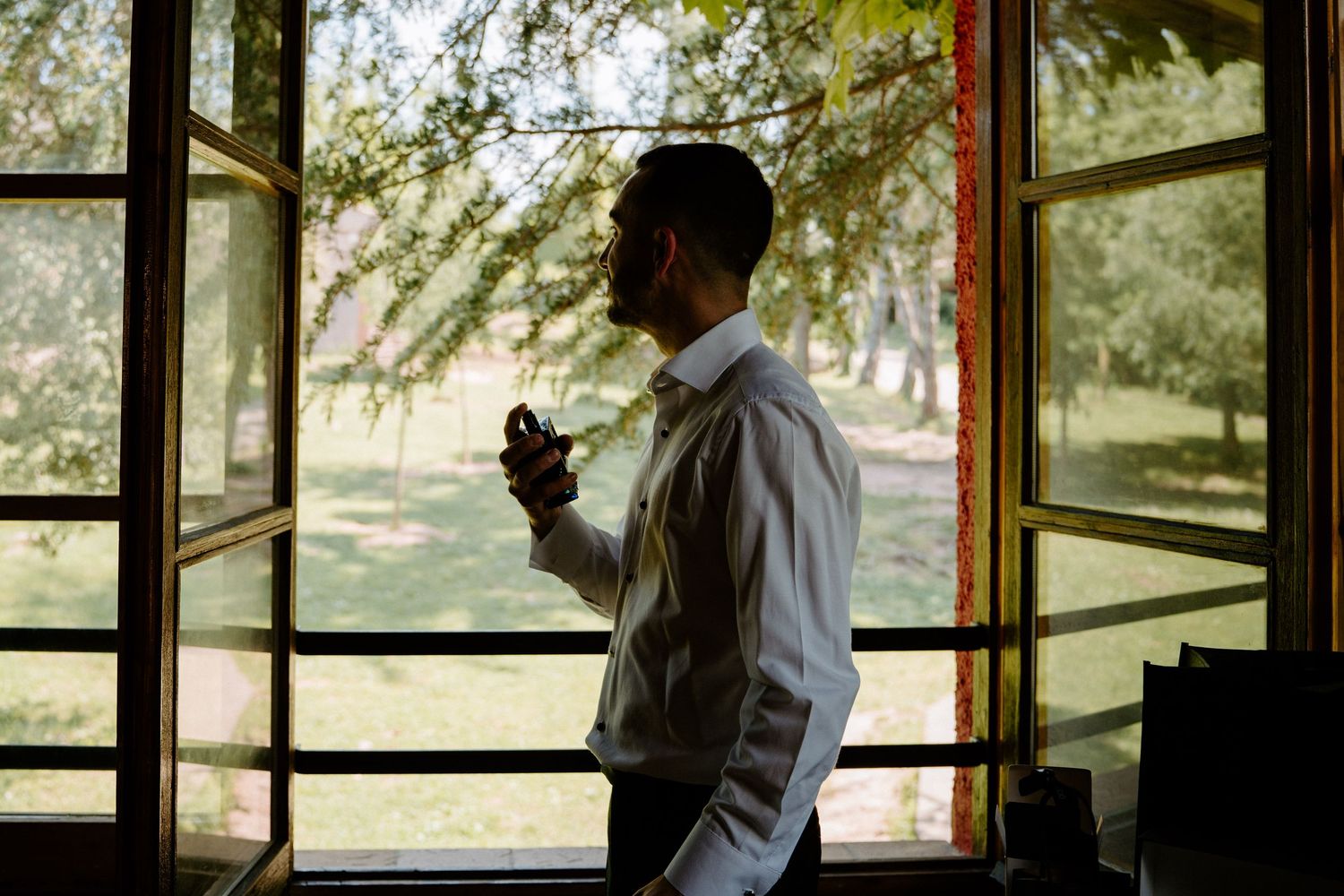 Groom in white shirt adjusting bow tie by large window with garden view in natural light.