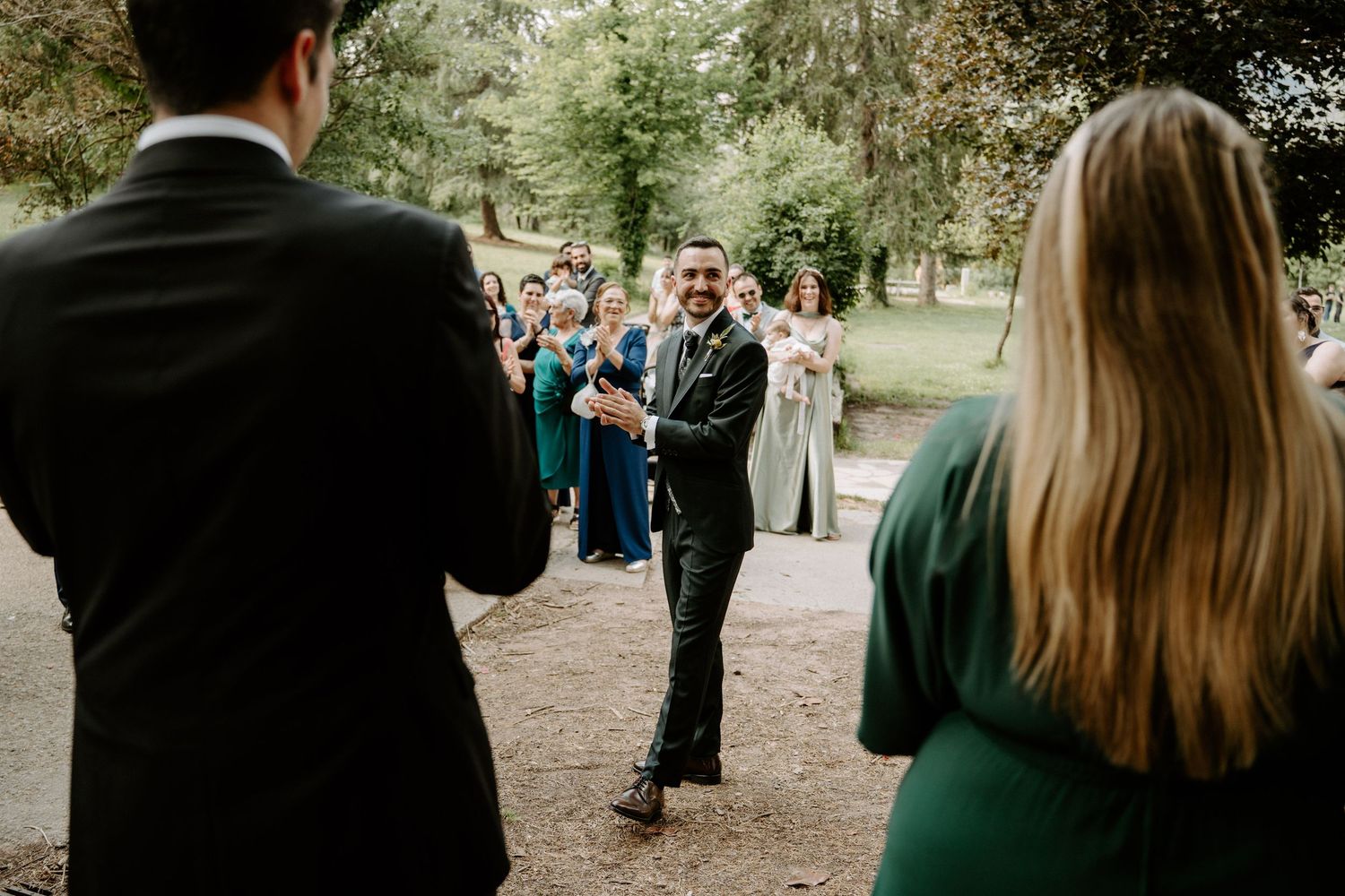 Smiling groom walks toward wedding party guests gathered along tree-lined outdoor pathway.