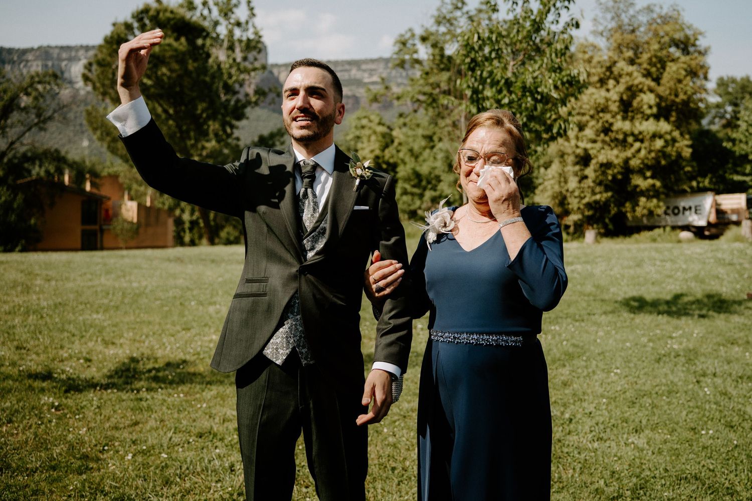 Groom in dark suit takes selfie with emotional mother wearing navy dress at outdoor wedding celebration.