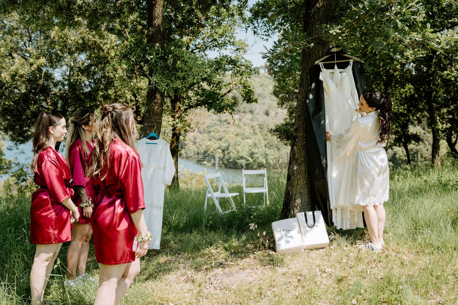 Bridesmaids in red robes admire wedding dress hanging from tree outdoors.