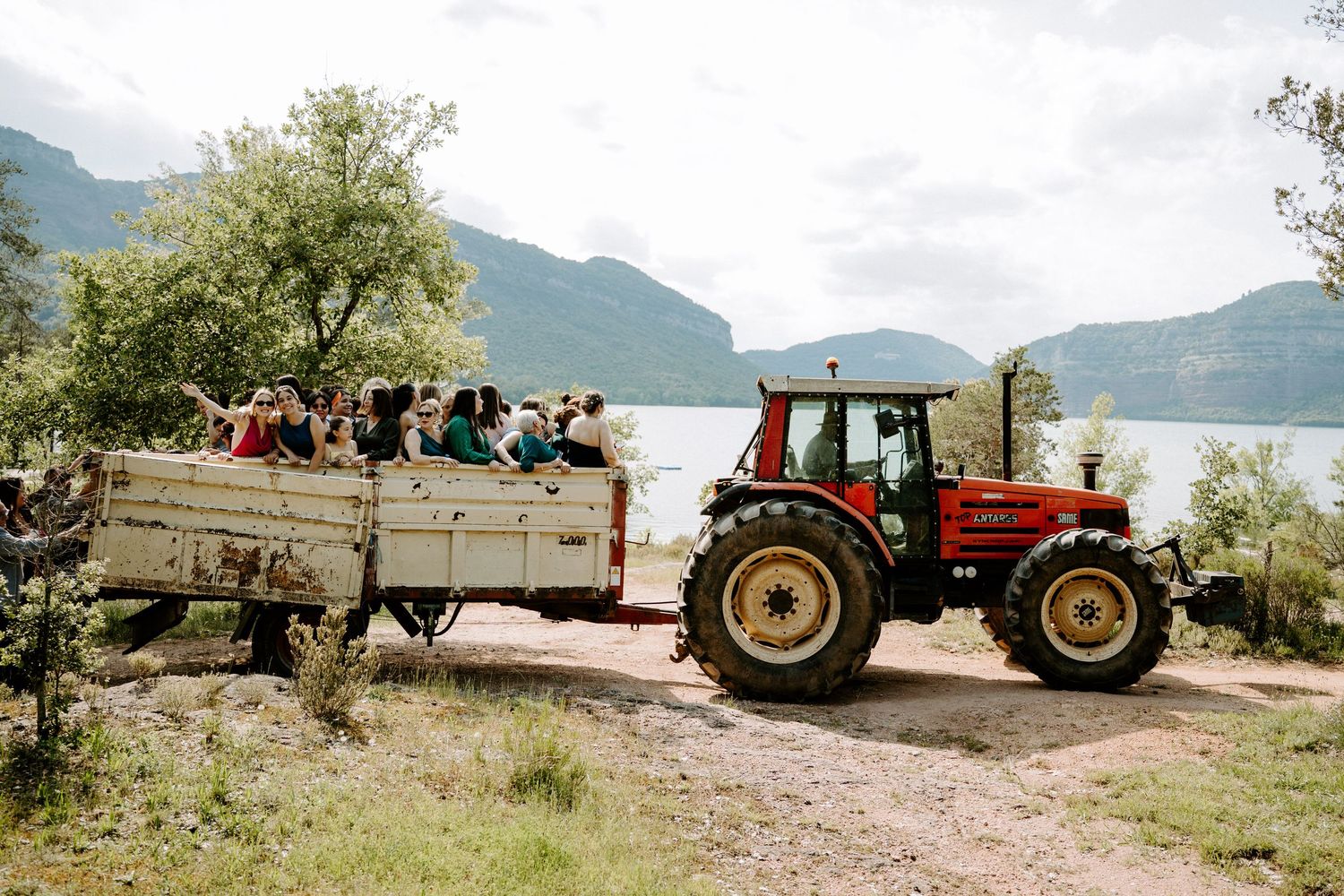 Wedding guests riding in a tractor-pulled trailer along a lakeside path with mountain views.