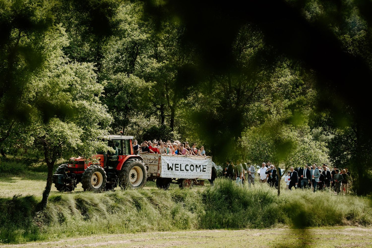 Red tractor pulls a welcome sign through lush green countryside at an outdoor wedding celebration.