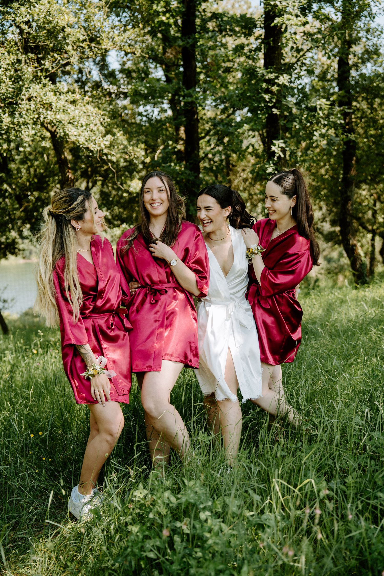 Bride and bridesmaids in red satin robes laughing together in a green forest meadow.
