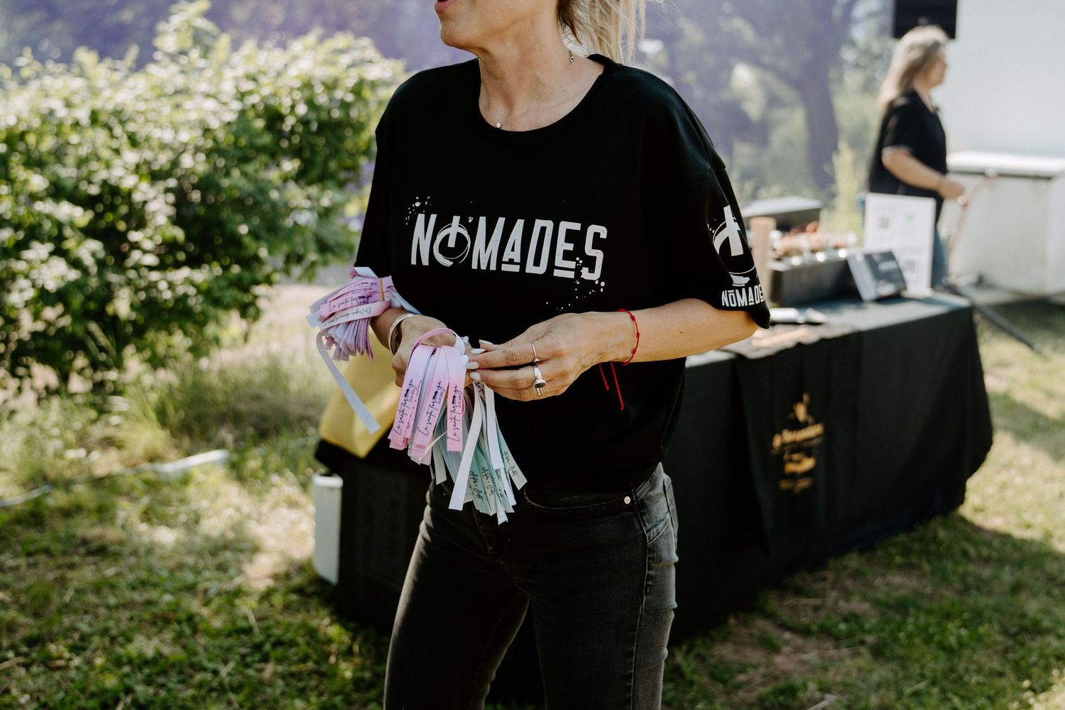 Woman in black Nomades t-shirt holding colorful ribbons at outdoor wedding event.