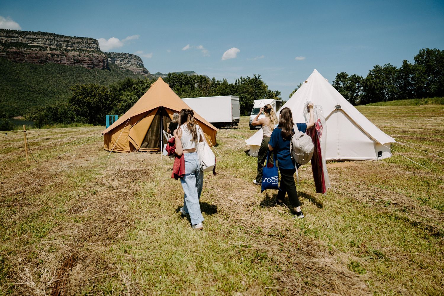 Wedding guests walking through a sunny field with bell tents and a mountain backdrop at an outdoor venue.