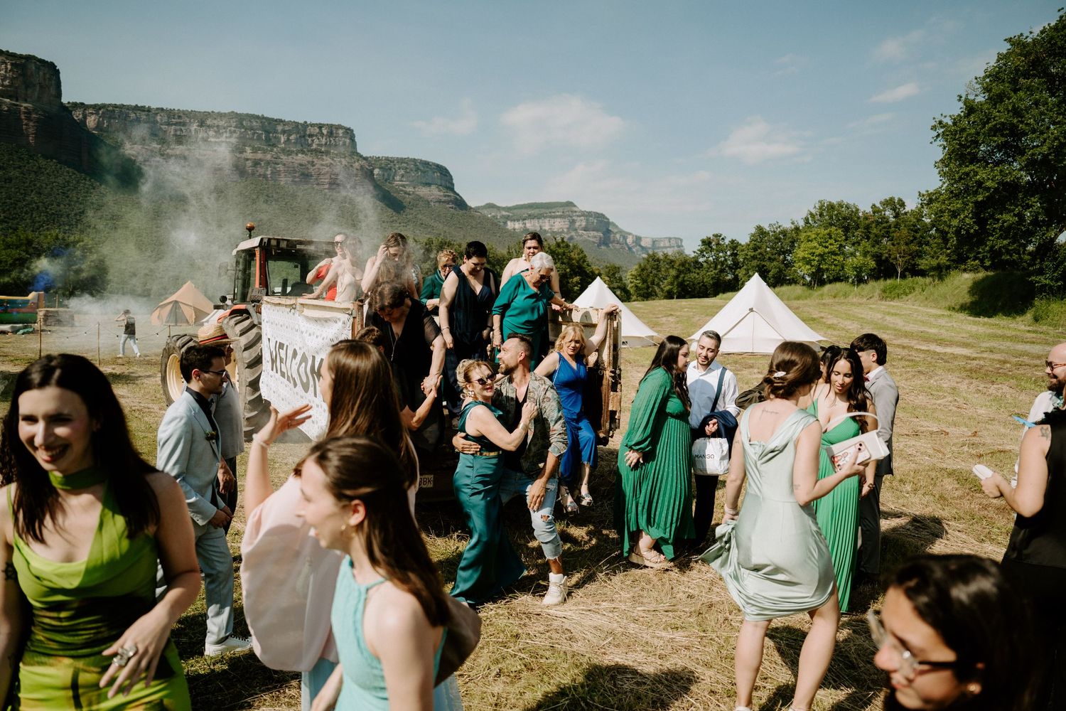 A lively outdoor wedding celebration with guests dancing and holding a sign in a scenic landscape.