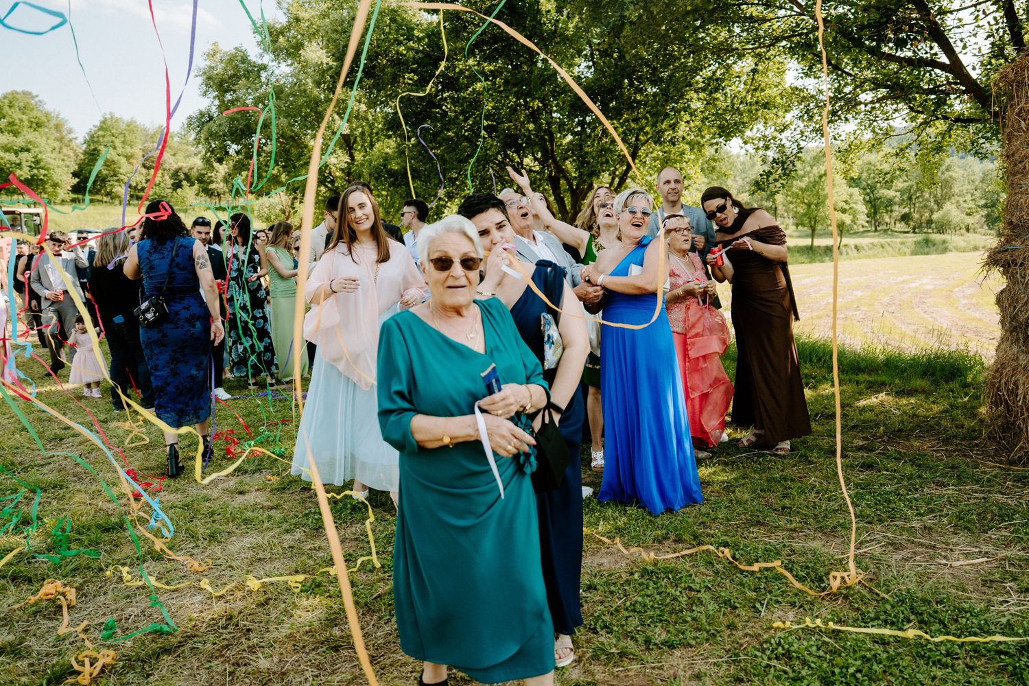 An elderly woman in teal leads wedding guests through a bamboo arch with colorful streamers.