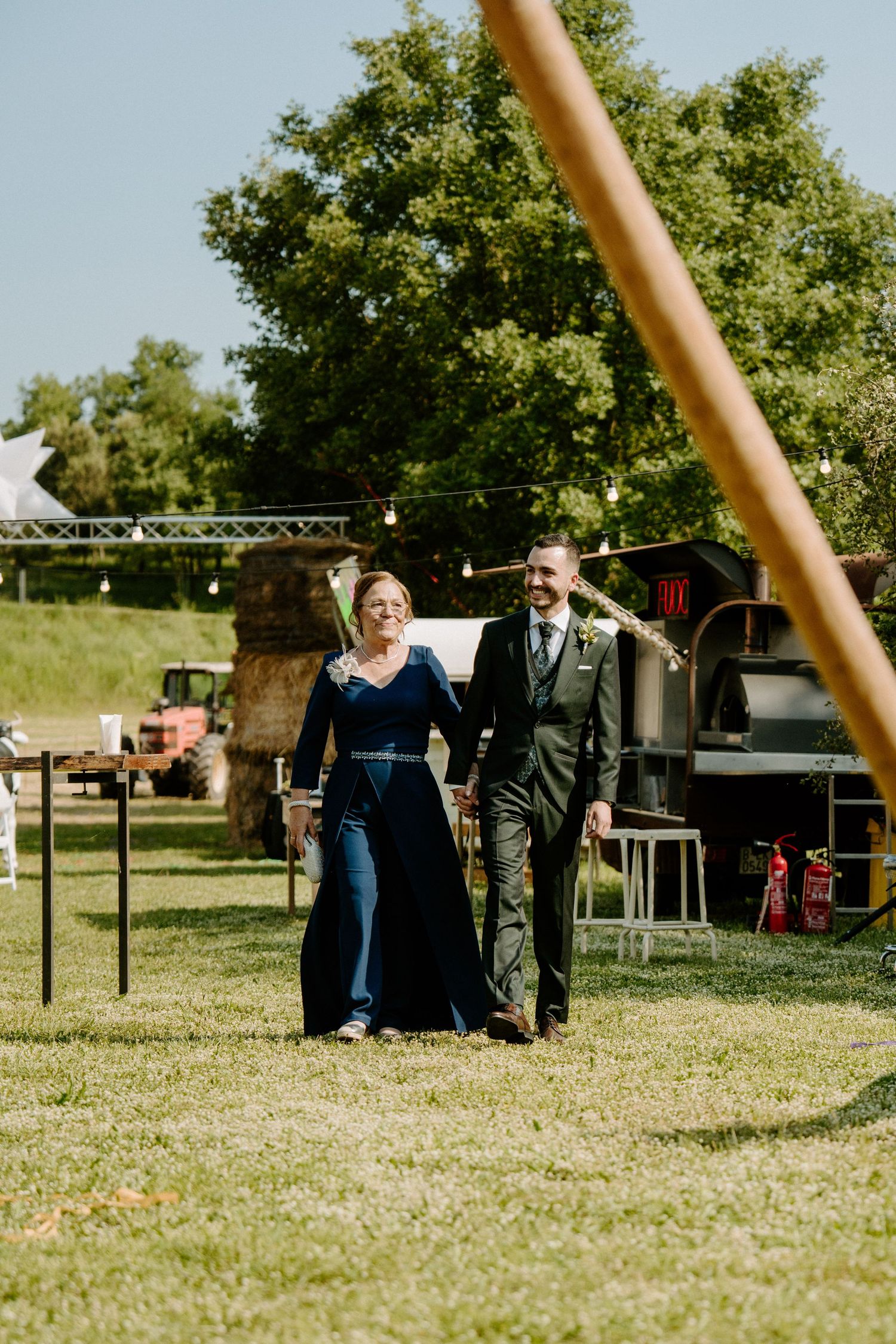 Elegant couple walks hand in hand across green lawn at rustic outdoor wedding venue.